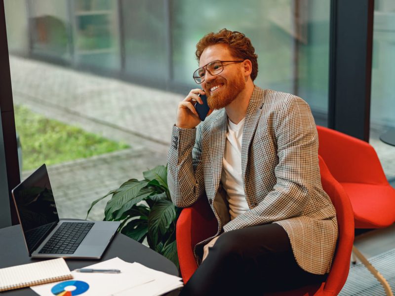 smiling-male-boss-talking-phone-with-client-while-sitting-in-coworking-space-and-working-on